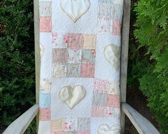 May include: A white quilt with a patchwork design featuring floral patterns in shades of pink, blue, and green. The quilt is decorated with four large, white satin hearts.