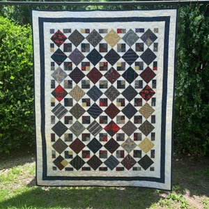 May include: A quilt made from a variety of patterned fabric squares in shades of blue, brown, red, and white. The squares are arranged in a diamond pattern and are sewn together with a white thread. The quilt is bordered with a white fabric and a dark blue fabric.