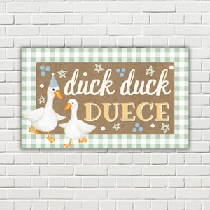 May include: A rectangular sign with a green and white checkered border and a brown center. The sign features two white ducks and the words "duck duck DUECE" in a playful font, with star and dot accents.