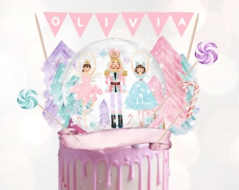 Puede incluir: Un pastel rosa con glaseado blanco y chispas rosas y blancas. El pastel está decorado con un adorno de bola de nieve con un cascanueces, una bailarina y un diseño de copo de nieve. El adorno también incluye una pancarta rosa con el nombre "OLIVIA" en ella.