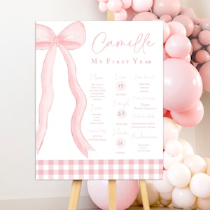 Puede incluir: Un fondo de cuadros vichy rosa y blanco con un lazo rosa y un cartel blanco que dice "Camille Mi Primer Año". El cartel enumera información sobre el primer año del niño, incluyendo su altura, peso, alimentos favoritos y apodos.