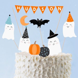 Pode incluir: Um bolo branco decorado com um tema de Halloween. O bolo é coberto com uma faixa que diz "HUDSON", um morcego, uma lua crescente e quatro fantasmas que usam chapéus de festa. Também há duas abóboras no bolo.