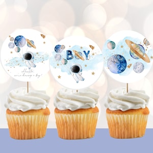 May include: Three cupcakes with white frosting topped with round paper toppers featuring a blue and gold astronaut holding balloons shaped like planets. The text "Houston, we're having a boy!" is on one topper, and the text "Boy" is on another topper.
