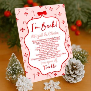 Puede incluir: Una tarjeta navideña rosa con el texto "I'm Back!" y un mensaje de un elfo llamado Twinkle. La tarjeta está decorada con bastones de caramelo y lazos, y está rodeada de decoraciones festivas.