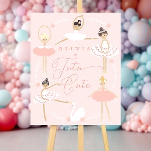 May include: A pink sign with a gold inscription that reads "Olivia is Tutu Cute". The sign is decorated with five ballerina illustrations wearing pink and white tutus. The sign is on a wooden easel.