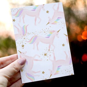 May include: A white card with a repeating pattern of pink unicorns with rainbow manes and tails. The background is white with gold glitter and stars.