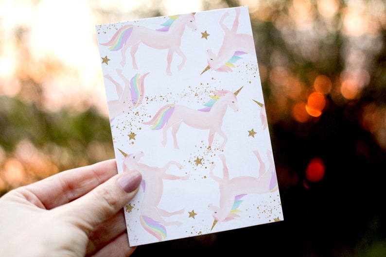 May include: A white card with a repeating pattern of pink unicorns with rainbow manes and tails. The background is white with gold glitter and stars.