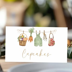 May include: A watercolor illustration on a white card features a clothesline with a basket of vegetables, carrots, green overalls, herbs, and a bag of apples. The word "Cupcakes" is written in cursive.