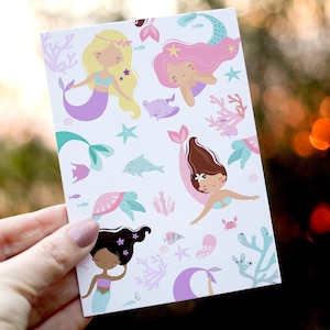 May include: A card with a white background and a repeating pattern of mermaids, sea turtles, starfish, and coral. The mermaids have long flowing hair in shades of pink, blonde, and brown. The mermaids are wearing seashell bras and tails with purple, pink, and green accents.
