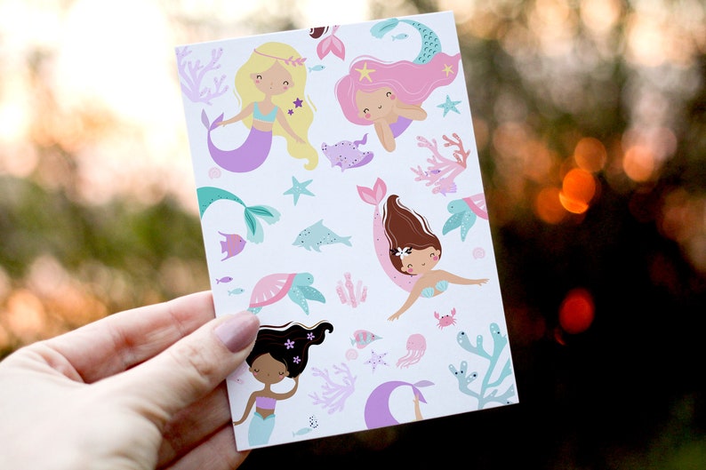 May include: A card with a white background and a repeating pattern of mermaids, sea turtles, starfish, and coral. The mermaids have long flowing hair in shades of pink, blonde, and brown. The mermaids are wearing seashell bras and tails with purple, pink, and green accents.