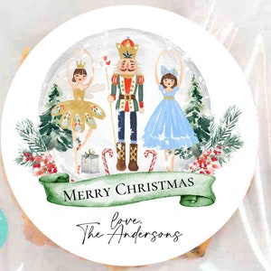 May include: A round Christmas sticker featuring a Nutcracker, a ballerina in a gold dress, and a ballerina in a blue dress. The sticker has a green banner with the text "Merry Christmas" and the text "love, The Andersons".