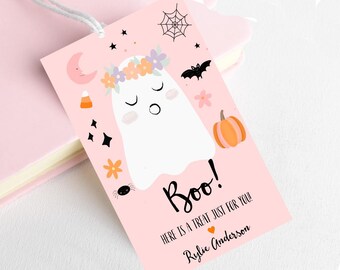 May include: A pink Halloween gift tag with a cartoon ghost wearing a floral crown. The tag says "Boo!" and "Here is a treat just for you!" with a name. Other graphics include a pumpkin, spider, and bat. The tag is tied with a white string.