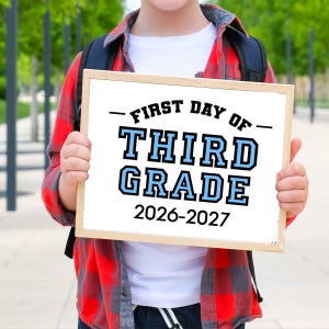May include: A rectangular wooden sign with white background and black and light blue text that reads FIRST DAY OF THIRD GRADE 2026-2027. The sign is held in front of a white t-shirt and a red and black plaid button-down shirt with a black backpack.