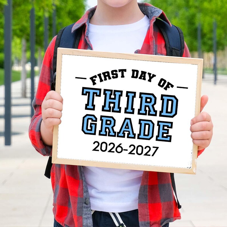 May include: A rectangular wooden sign with white background and black and light blue text that reads FIRST DAY OF THIRD GRADE 2026-2027. The sign is held in front of a white t-shirt and a red and black plaid button-down shirt with a black backpack.