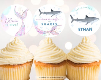 May include: Three cupcake toppers with a mermaid and shark theme. The first topper says "Olivia is Five!" and features a mermaid tail with pink, purple, and blue scales. The second topper says "mermaids and sharks" and features a mermaid tail with gold glitter. The third topper says "Happy Birthday Ethan" and features a gray shark with white bubbles.