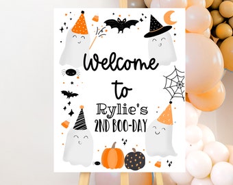 May include: A white sign with black and orange Halloween decorations, including ghosts, bats, spiders, and pumpkins. The sign says "Welcome to Rylie's 2nd Boo-Day".