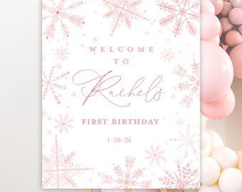 May include: A white sign with pink snowflake designs and the text "WELCOME TO Rachel's FIRST BIRTHDAY 1-20-26". The sign is displayed on a wooden easel, with pink balloons in the background.