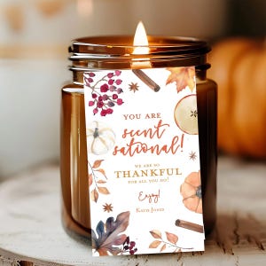 May include: A lit brown glass jar candle with a decorative tag. The tag features watercolor illustrations of pumpkins, leaves, and berries, with the text "YOU ARE scent-sational!" and "WE ARE SO THANKFUL".