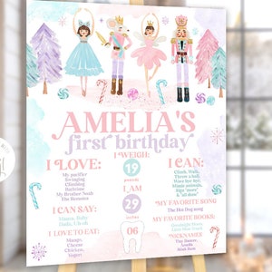 May include: A pink and blue first birthday sign with a watercolor background featuring a ballerina, a nutcracker, and a mouse. The sign includes the child's name, Amelia, and details about her first year, including her weight, height, and favorite things.