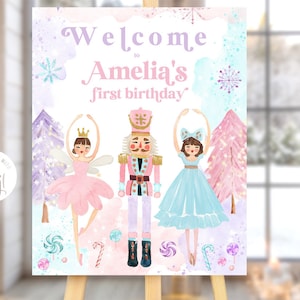 May include: A welcome sign for a first birthday party with a Nutcracker theme. The sign features a pink, blue, and purple watercolor background with snowflakes and candy canes. The text "Welcome to Amelia's first birthday" is written in pink and purple lettering. There are three figures on the sign: a ballerina in a pink tutu, a Nutcracker soldier, and a ballerina in a blue tutu.