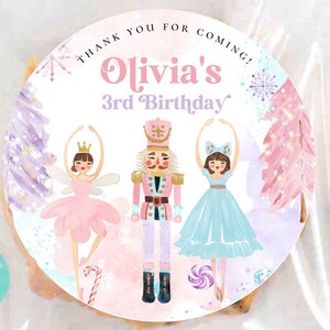 May include: A round sticker featuring a nutcracker flanked by two ballerinas in pink and blue dresses. The text reads "THANK YOU FOR COMING!" and "Olivia's 3rd Birthday". The design includes watercolor trees, snowflakes, and candy canes in soft pastel colors.