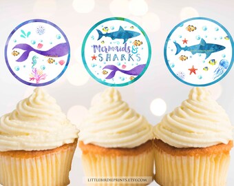 May include: Three cupcakes topped with round, colorful cupcake toppers featuring mermaids and sharks. The toppers have a watercolor design with blue, purple, and green colors. The text on one topper reads "Mermaids & Sharks".