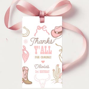 Könnte beinhalten: Ein weißes Geschenk-Etikett mit rosa Cowboy-Illustrationen und Text. Auf dem Etikett steht "Thanks Y'all for Coming!" und enthält den Namen "Olivia's" und "3rd Birthday". Oben ist ein rosa Band gebunden.