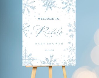 Puede incluir: Un letrero rectangular blanco con ilustraciones de copos de nieve en azul claro a lo largo de los bordes. El texto dice: WELCOME TO Rachel's BABY SHOWER 01-14-26. El letrero se exhibe en un caballete de madera sobre un fondo azul.