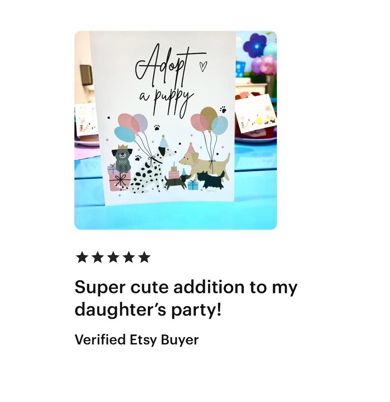 May include: A white card with the text "Adopt a puppy" in black script with a small pink heart. The card features a colorful illustration of four dogs with party hats and balloons. The card is on a blue table with a pink and white tablecloth.