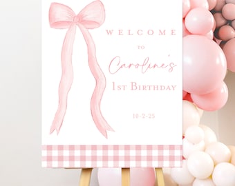 May include: A pink and white welcome sign with a large pink bow and the text "Welcome to Caroline's 1st Birthday 10-2-25". The sign is on a wooden easel and has a pink and white gingham pattern at the bottom.