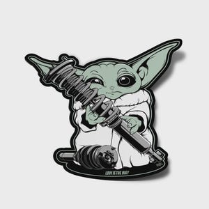 Puede incluir: Una pegatina con un personaje de dibujos animados con orejas grandes, sosteniendo piezas de suspensión de coche. El personaje es verde y lleva una túnica blanca. La pegatina tiene un contorno negro y el texto "LOW IS THE WAY" en la parte inferior.