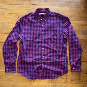 May include: A long-sleeved button-down shirt in a classic gingham pattern. The shirt features a dark red and navy blue checkered design, a button-down collar, and white buttons. The shirt is made of a woven fabric.