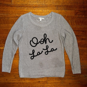 Peut inclure: Un pull en tricot gris avec des manches longues et un col rond. L'expression "Ooh La La" est brodée en noir sur le devant. Le pull est fait d'un tissu tricoté texturé.