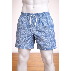 Könnte beinhalten: Blaue Badeshorts mit weißem Ananasmuster. Die Shorts haben einen Kordelzug in der Taille und werden auf einer Schaufensterpuppe präsentiert. Die knielangen Shorts eignen sich für Strand oder Pool.