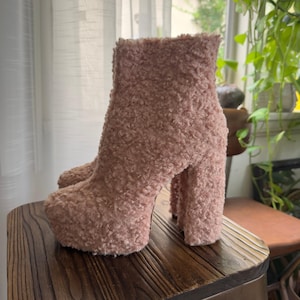May include: A pair of pink, fluffy ankle boots with a platform sole and a chunky heel. The boots are made of a soft, textured material and sit on a wooden surface. The boots have a height of approximately 6 inches.