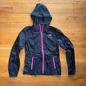 May include: Black hooded jacket with a bright pink zip and pocket details. The jacket features a drawstring hood and the brand logo is visible on the chest. The jacket is lightweight and appears to be made of a water-resistant material.