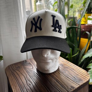 May include: A baseball cap with a white crown and a black brim. The cap features the navy blue letters "NY" and "LA" on the front. The hat is displayed on a white mannequin head.