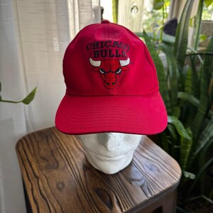 Peut inclure: Casquette de baseball rouge avec le logo des Chicago Bulls brodé sur le devant. Le logo comprend le nom de l'équipe et une tête de taureau. La casquette a une visière incurvée et une couronne structurée.