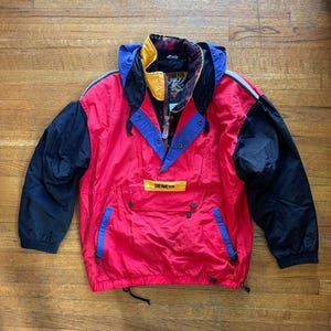 May include: A red, black, and blue Obermeyer anorak jacket. The jacket has a hood, a half-zip closure, and two front pockets. The sleeves are black, and the body is red with blue and yellow accents. The jacket has the Obermeyer logo on the front.