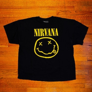 Peut inclure: T-shirt noir avec le mot "NIRVANA" en lettres jaunes au-dessus d'un smiley jaune avec des yeux barrés et une langue tirée. Le t-shirt est fait d'une matière douce.