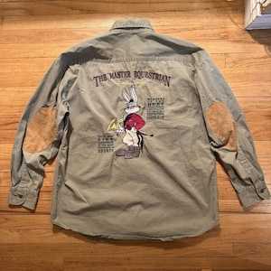 May include: A long-sleeved, olive green button-down shirt with brown elbow patches. The back features a Bugs Bunny cartoon graphic with the text "THE MASTER EQUESTRIAN" and other text. The shirt has a collared neckline and buttoned cuffs.