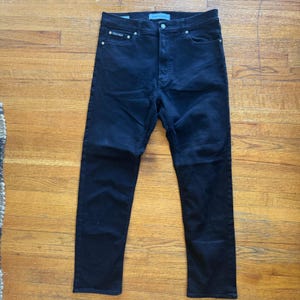 Puede incluir: Vaqueros de mezclilla azul marino oscuro con cierre de botón y cremallera. Los vaqueros tienen un diseño clásico de cinco bolsillos y un logotipo de Calvin Klein en el bolsillo delantero. Los vaqueros están planos sobre una superficie de madera.