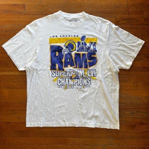 May include: A white short-sleeved t-shirt featuring a graphic print for the Los Angeles Rams Super Bowl LVI Champions. The design includes the team name, a football helmet, the Vince Lombardi trophy, and the text Los Angeles, California February 13, 2022.