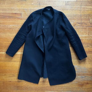 May include: A black long coat with a draped collar and long sleeves. The coat is made of a soft material and has a single button closure. The coat is laid out on a wooden surface.