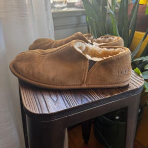 May include: A pair of brown suede slippers with a sheepskin lining. The slippers are slip-on style and have a stitched sole. The brand name "UGG" is visible on the side of one slipper. The slippers are resting on a small wooden stool.