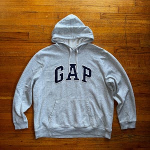 May include: A heather gray hooded sweatshirt with the word "GAP" in navy blue block letters across the chest. The sweatshirt has a front pocket and a drawstring hood. The garment is made of a soft, comfortable material.