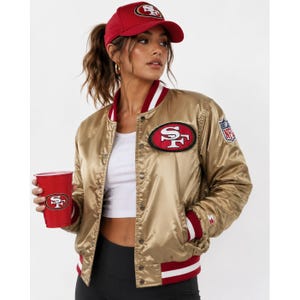 May include: A gold satin bomber jacket with red and white striped trim, featuring a San Francisco 49ers logo patch on the chest and an NFL shield on the sleeve. The jacket is worn with a red baseball cap with a 49ers logo and a red plastic cup.