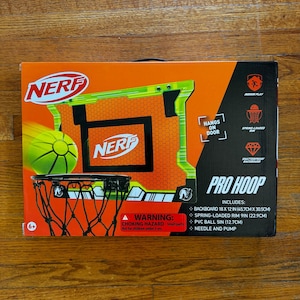May include: Nerf Pro Hoop set in a box. The box is orange and black with the Nerf logo and a basketball graphic. Includes a backboard (18 x 12 in), spring-loaded rim (9 in), PVC ball (5 in), and needle and pump.