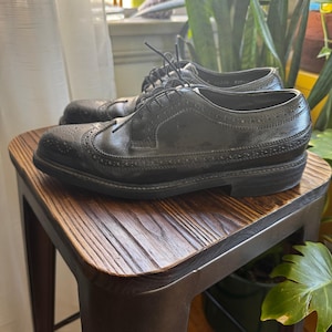 May include: A pair of black leather dress shoes with brogue detailing. The shoes have a classic wingtip design with decorative perforations and are laced up. They are placed on a wooden stool.