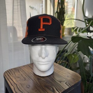 May include: Black and orange baseball cap with the letter "P" in orange on the front. The cap has a flat brim and a structured crown. The cap is displayed on a white mannequin head.